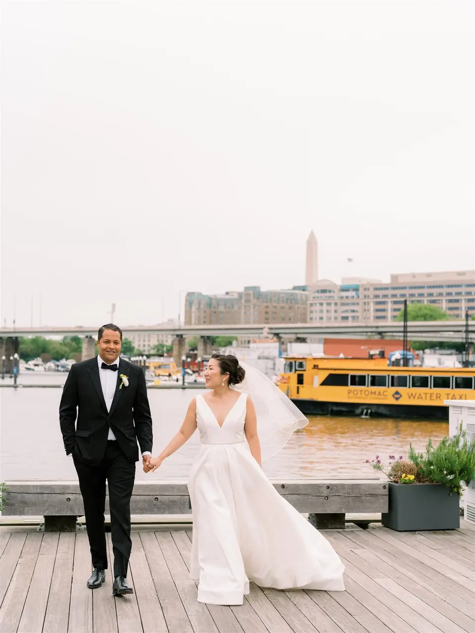 Bride and groom walking hand in hand on a boardwalk beside the water with a yellow water taxi behind