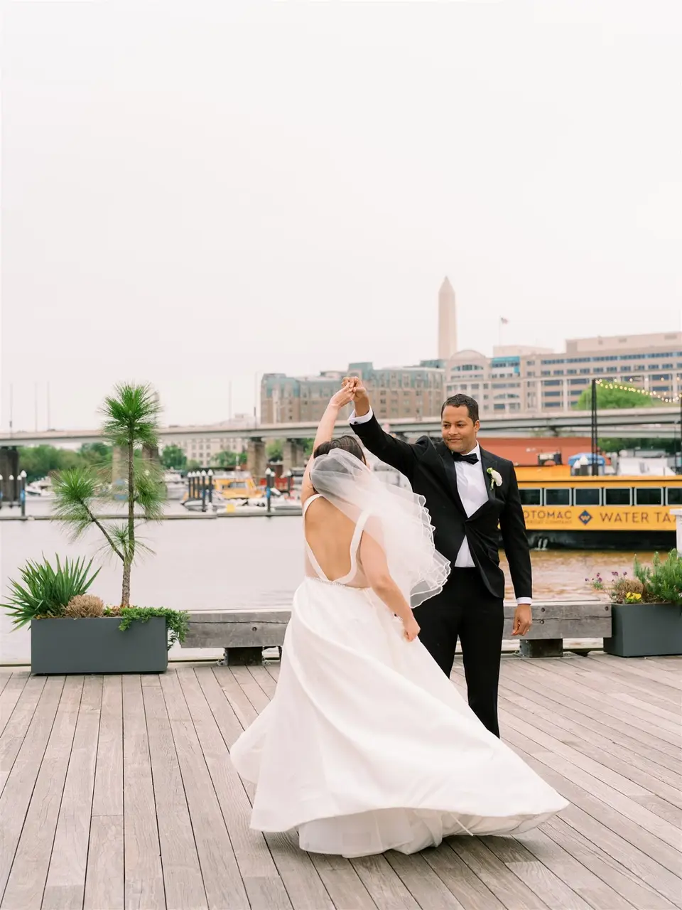 Groom twirling the bride on a waterfront boardwalk with a water taxi and Washington Monument behind