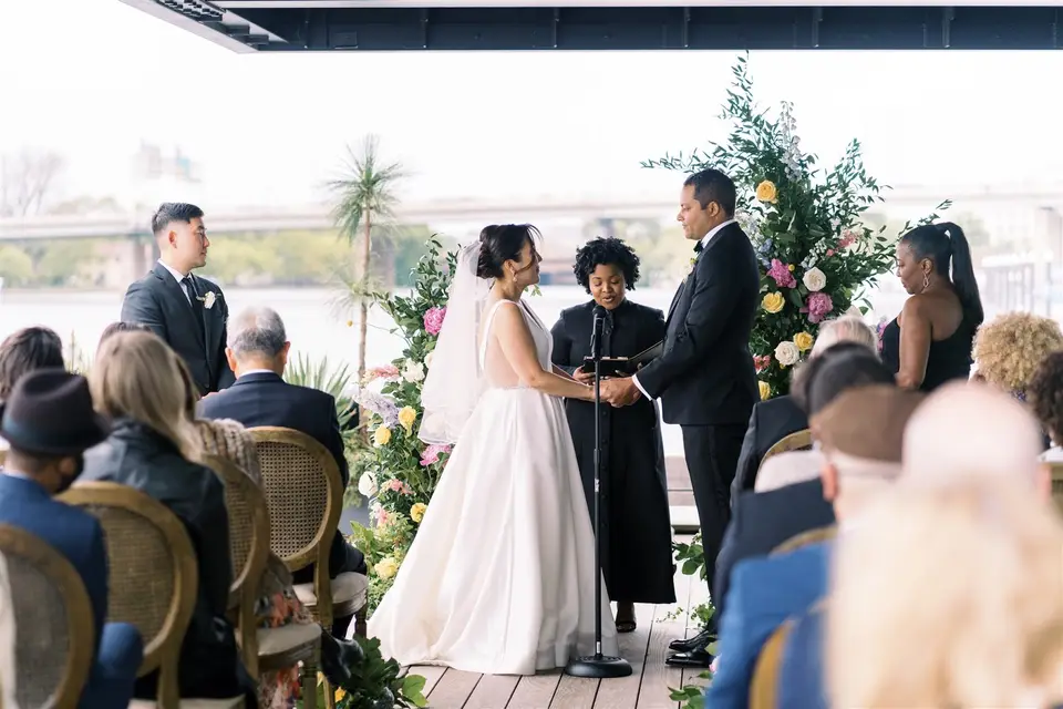 Bride and groom holding hands facing the officiant at a waterfront ceremony with colorful flowers and seated guests