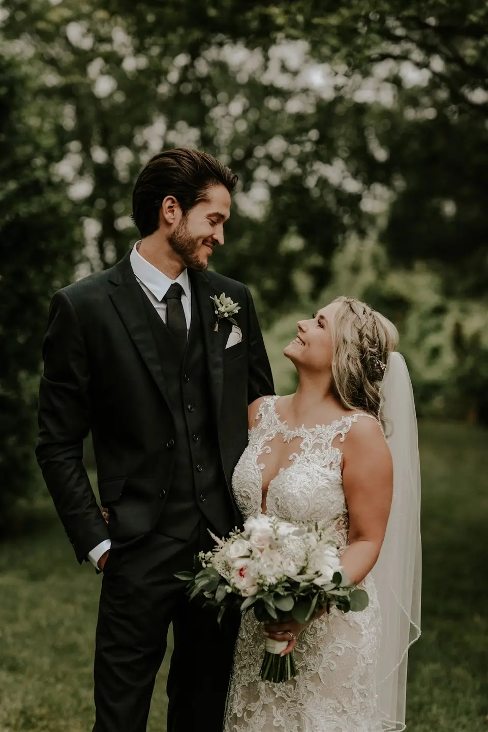 Bride in a lace gown and veil smiles up at the groom in a dark suit as they pose outdoors in a green garden