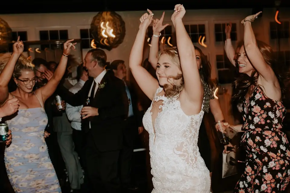 Smiling bride in a lace gown dances with her arms raised among guests holding drinks