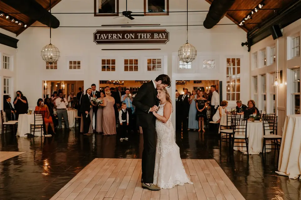 Bride and groom dance on a light wood dance floor under a Tavern on the Bay sign as guests look on