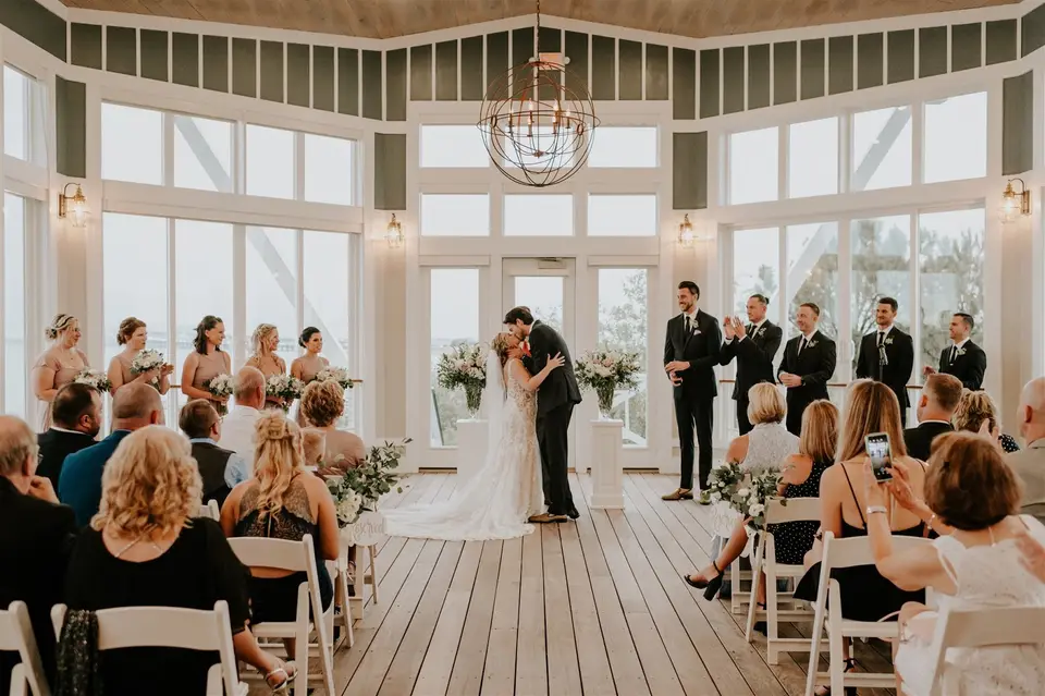 Bride and groom sharing a kiss during a ceremony in a glass-walled room with guests seated and groomsmen standing