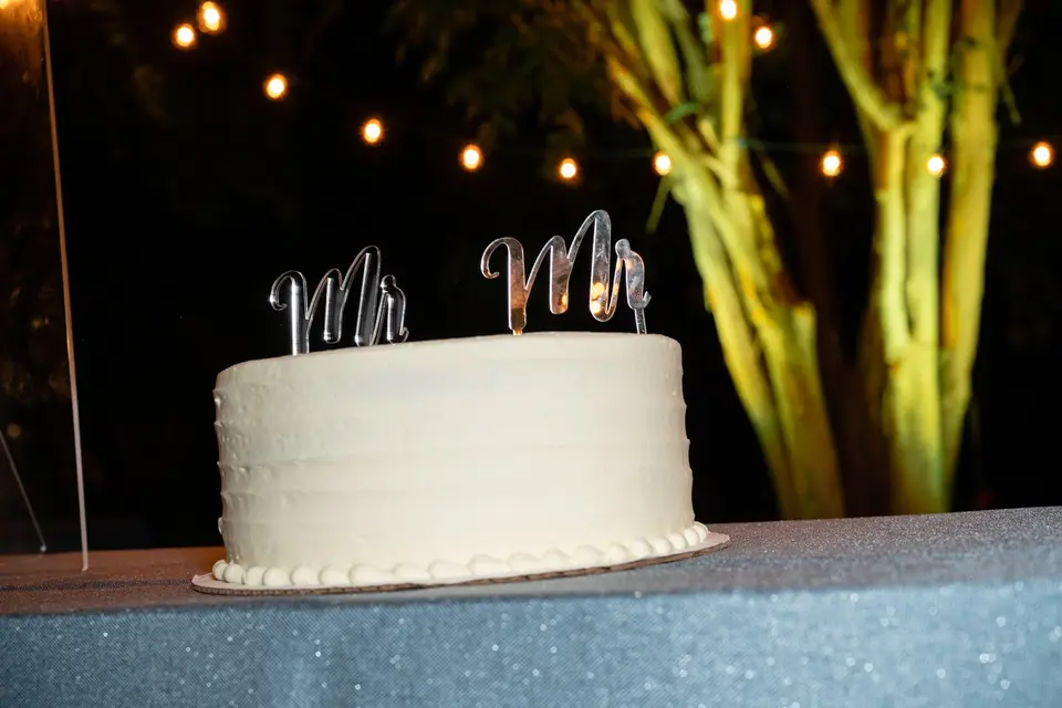 White wedding cake topped with silver Mr and Mr letters on a sparkly table at an outdoor reception