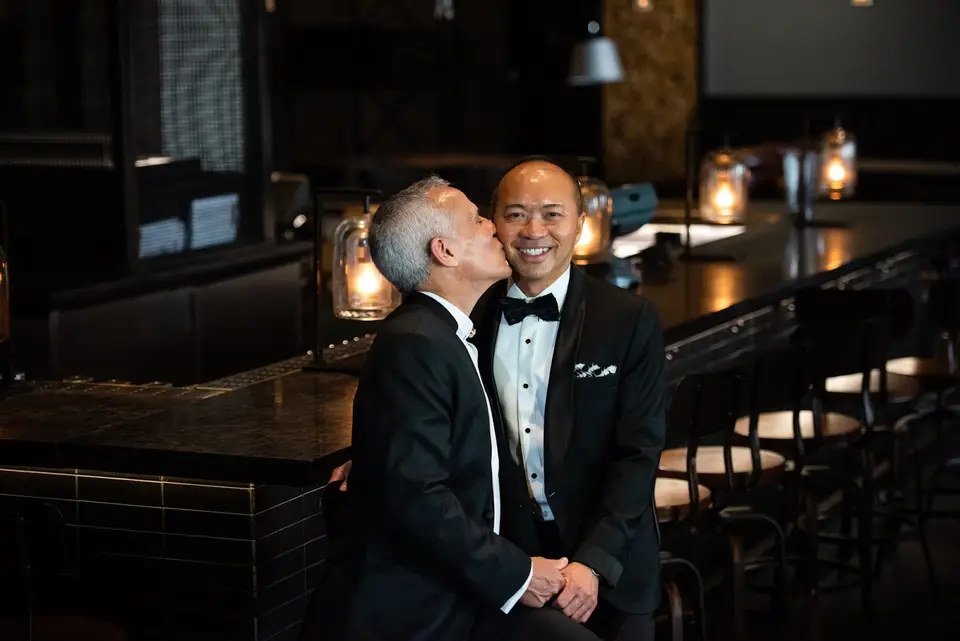 Groom in a tuxedo kissing his husband on the cheek at a dark bar