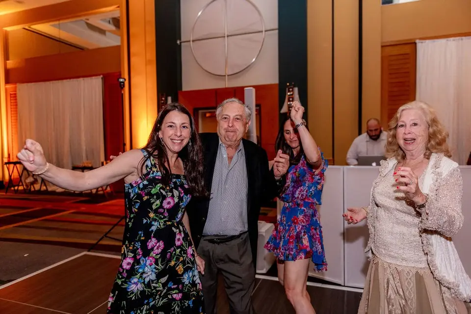 Three guests dancing and laughing near the DJ table at a hotel ballroom reception