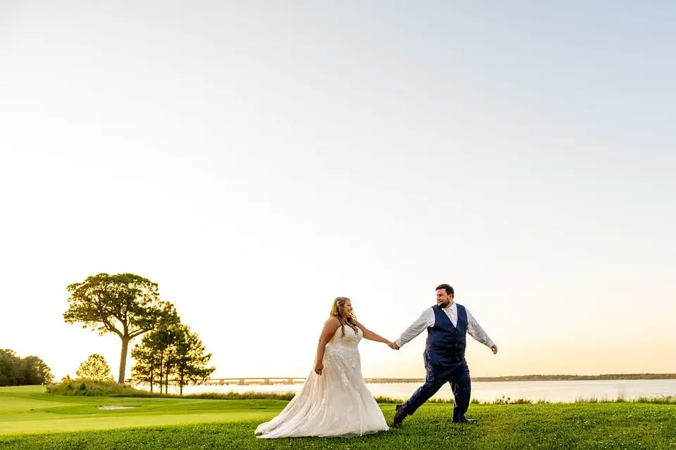 Bride and groom holding hands and walking across a waterfront golf course lawn at sunset