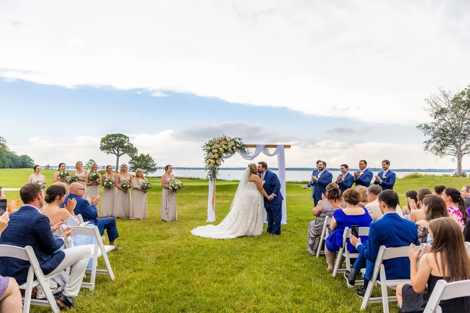 Bride and groom kissing under a floral arch on a lawn by the water as guests and wedding party look on