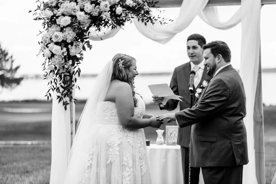 Black and white photo of the bride and groom holding hands under a floral arch as the officiant reads