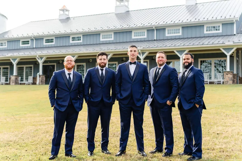 Five groomsmen in navy suits standing on grass in front of a long barn building