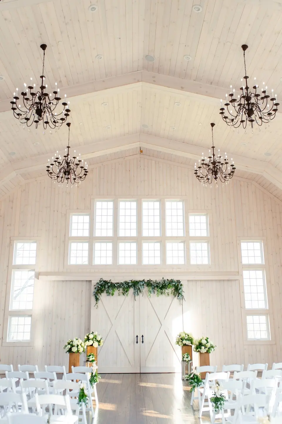 Wedding ceremony space inside a white barn with chandeliers, a greenery-topped barn door and rows of white chairs