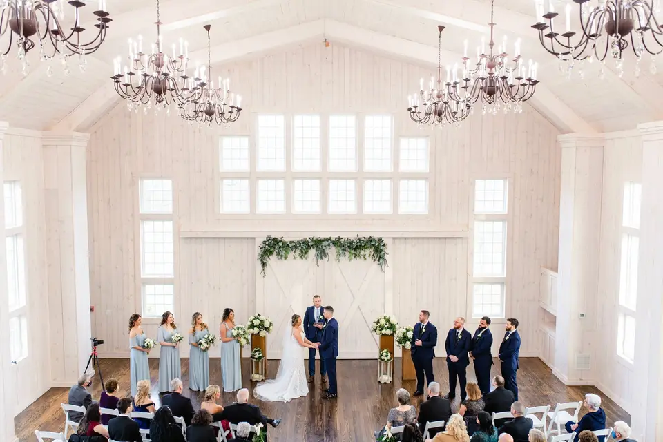 Overhead view of a wedding ceremony inside a white barn venue with chandeliers and guests seated