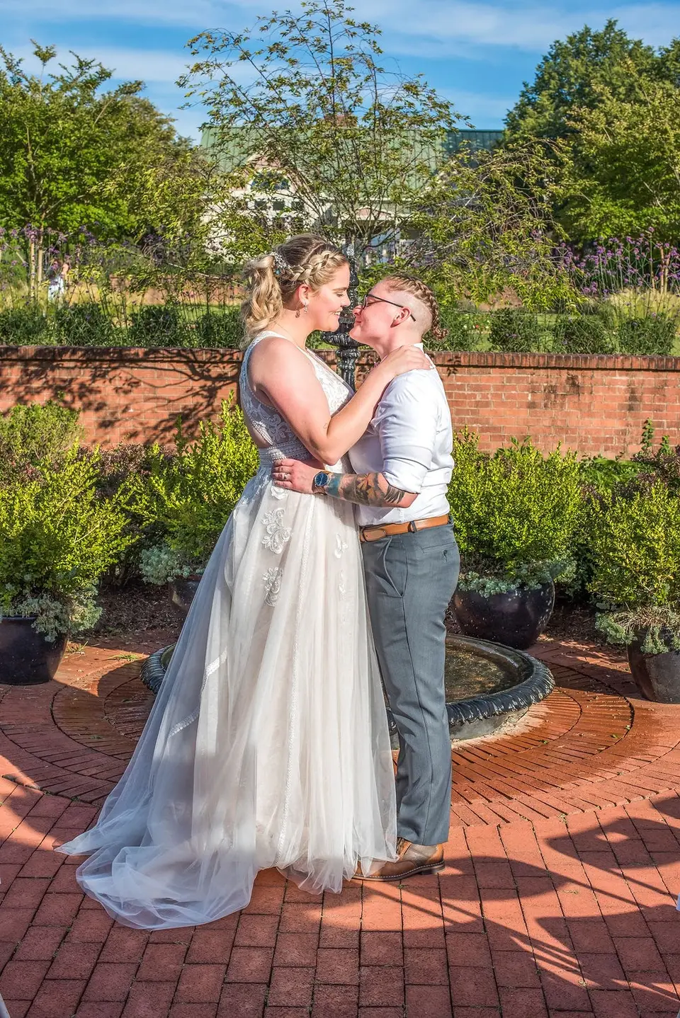 Two brides embracing on a brick garden patio with a fountain and planters behind them