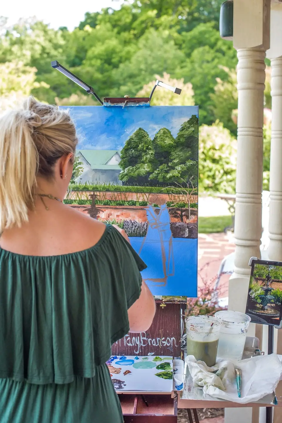 Close view of a woman painting a garden scene on an easel at a wedding, with paint jars on a small table