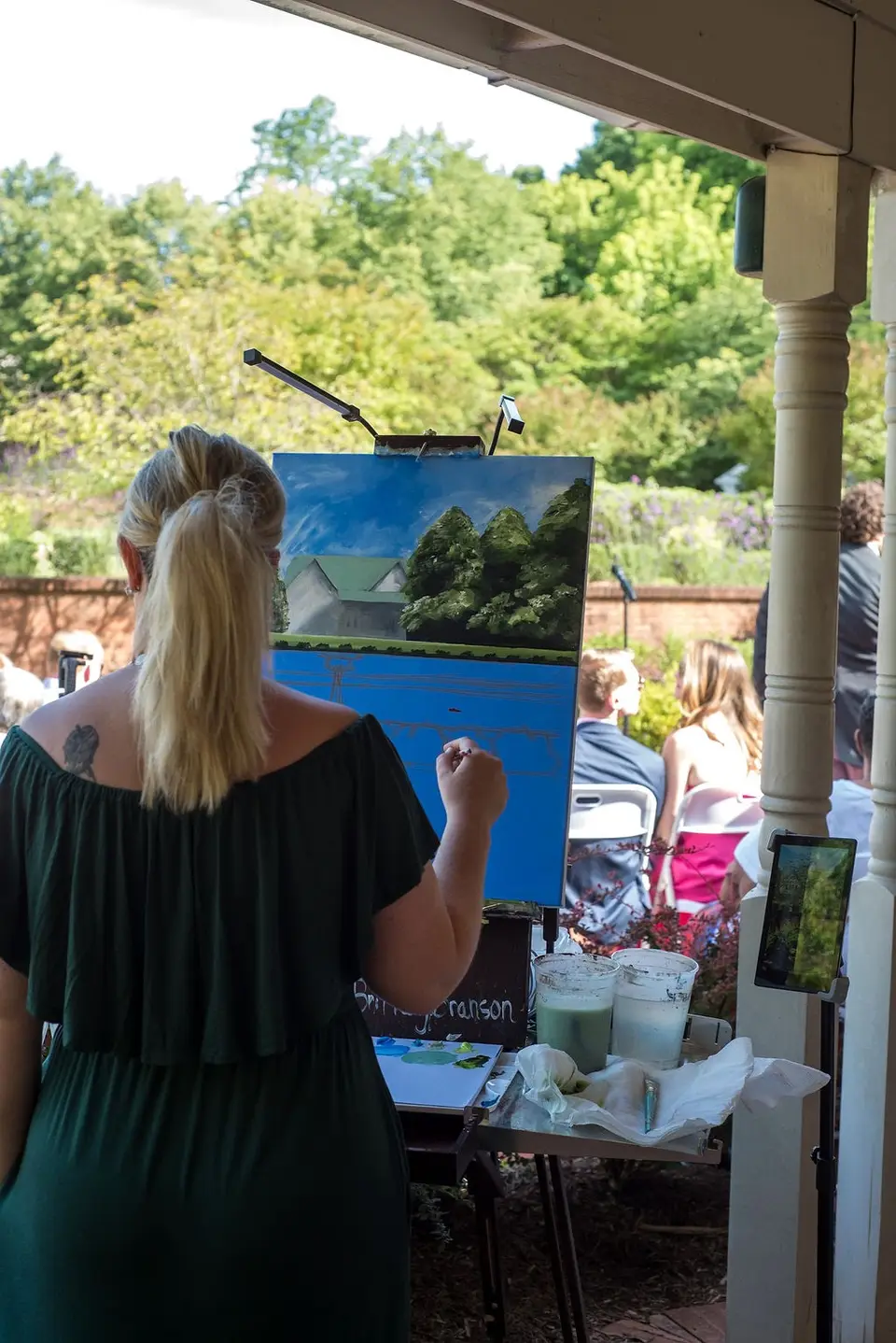 Woman in a green off-the-shoulder dress paints a landscape on an easel on a porch during a wedding