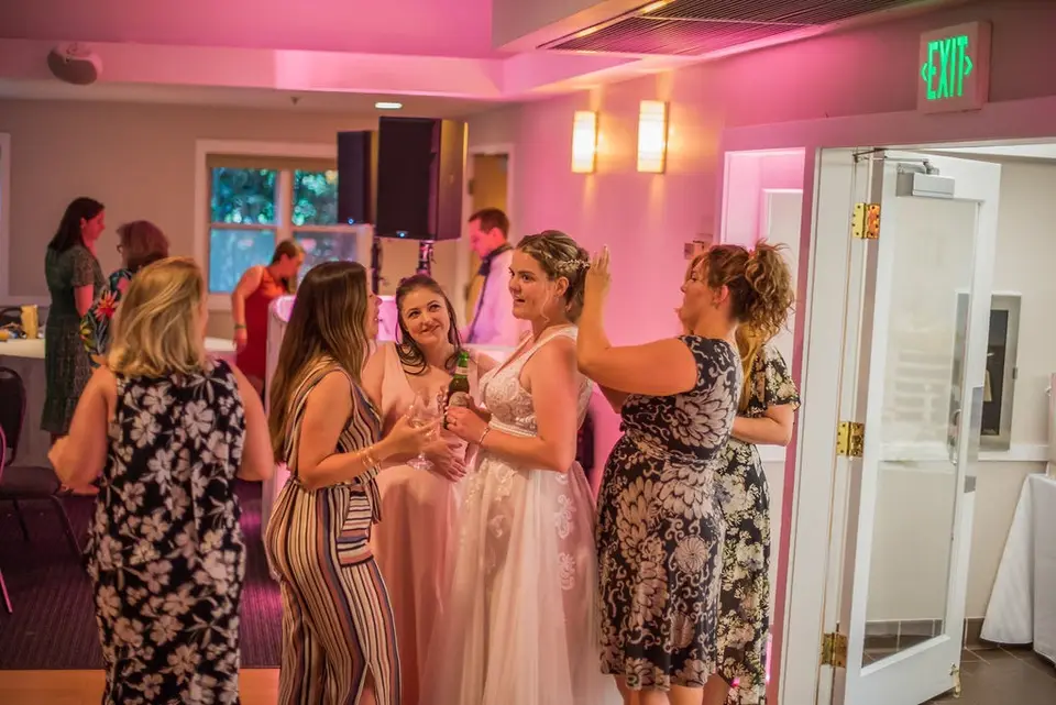 Bride in a white gown talks and holds a drink with female guests in a pink-lit room near an exit sign