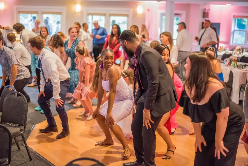Wedding guests dancing and laughing on a wooden dance floor in a pink-lit reception room