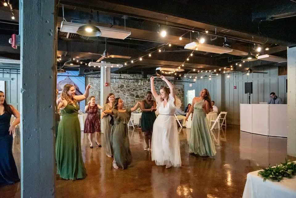 Bride and bridesmaids in green dresses dancing together on a polished floor under string lights