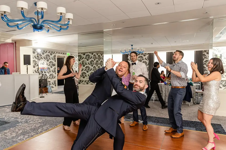 Groom dipping his partner backward on the dance floor as guests dance and cheer in a hotel ballroom