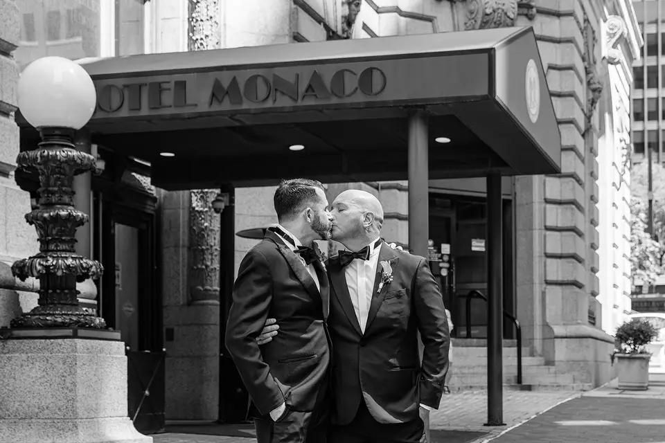 Two grooms in tuxedos kissing under the Hotel Monaco entrance canopy in black and white
