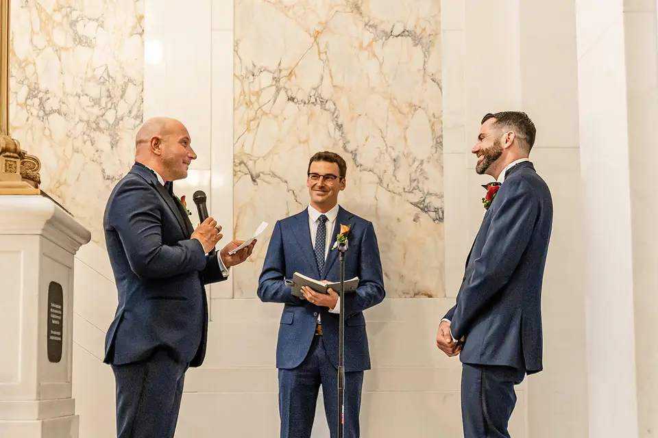 Officiant holding a book between two grooms as one speaks into a microphone during a marble hall ceremony