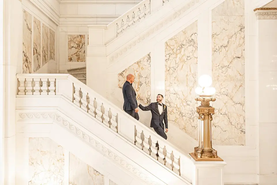 Two grooms in dark suits climbing a white marble staircase with a gold lamp post