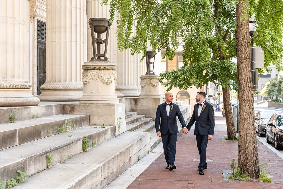 Two grooms in navy tuxedos holding hands and walking down a tree-lined brick sidewalk