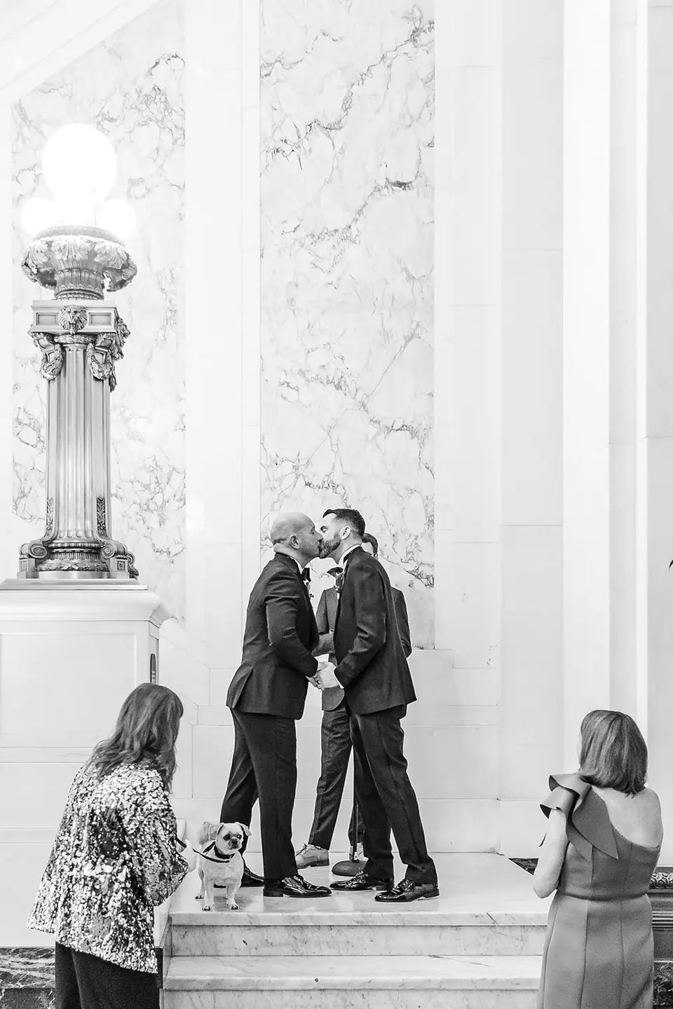Black and white photo of two grooms kissing at the altar as a small dog and guests look on