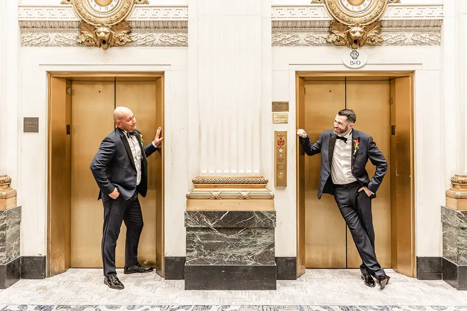 Two grooms in tuxedos leaning against separate gold elevator doors in an ornate lobby