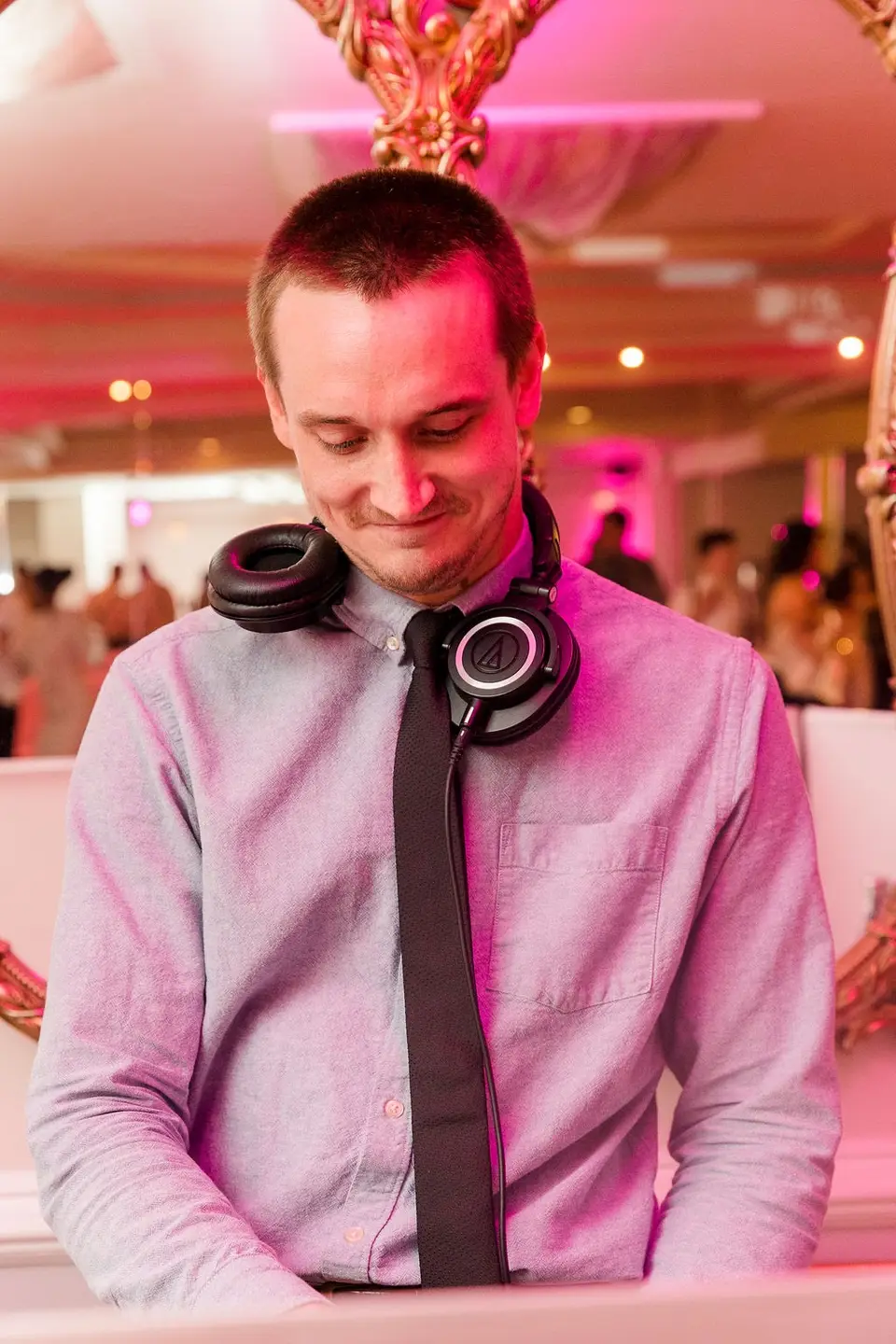 DJ in headphones draped around his neck smiling down at his equipment under pink lighting