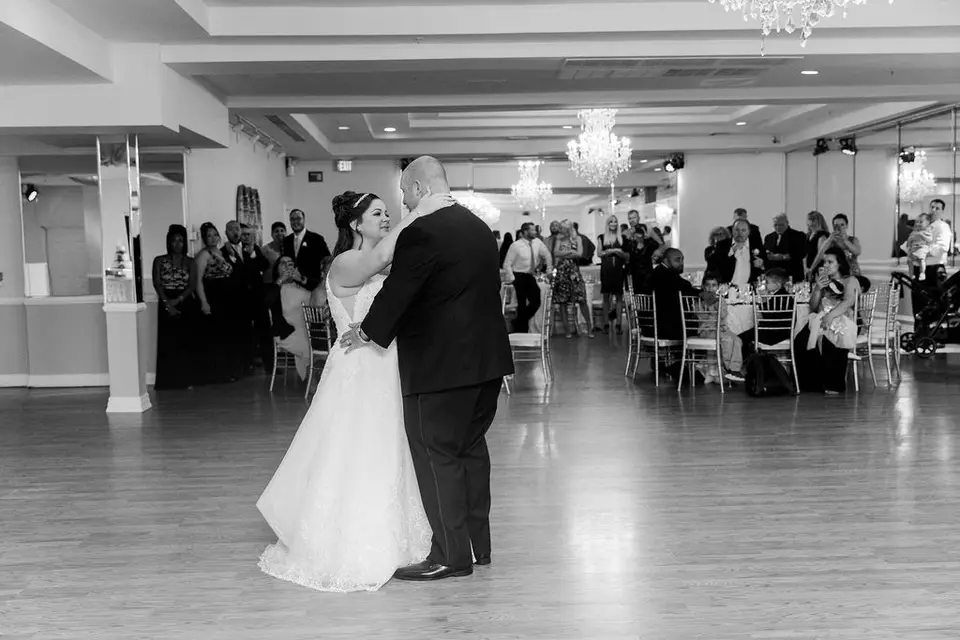 Black and white photo of the bride and groom sharing a first dance in a banquet hall as guests watch