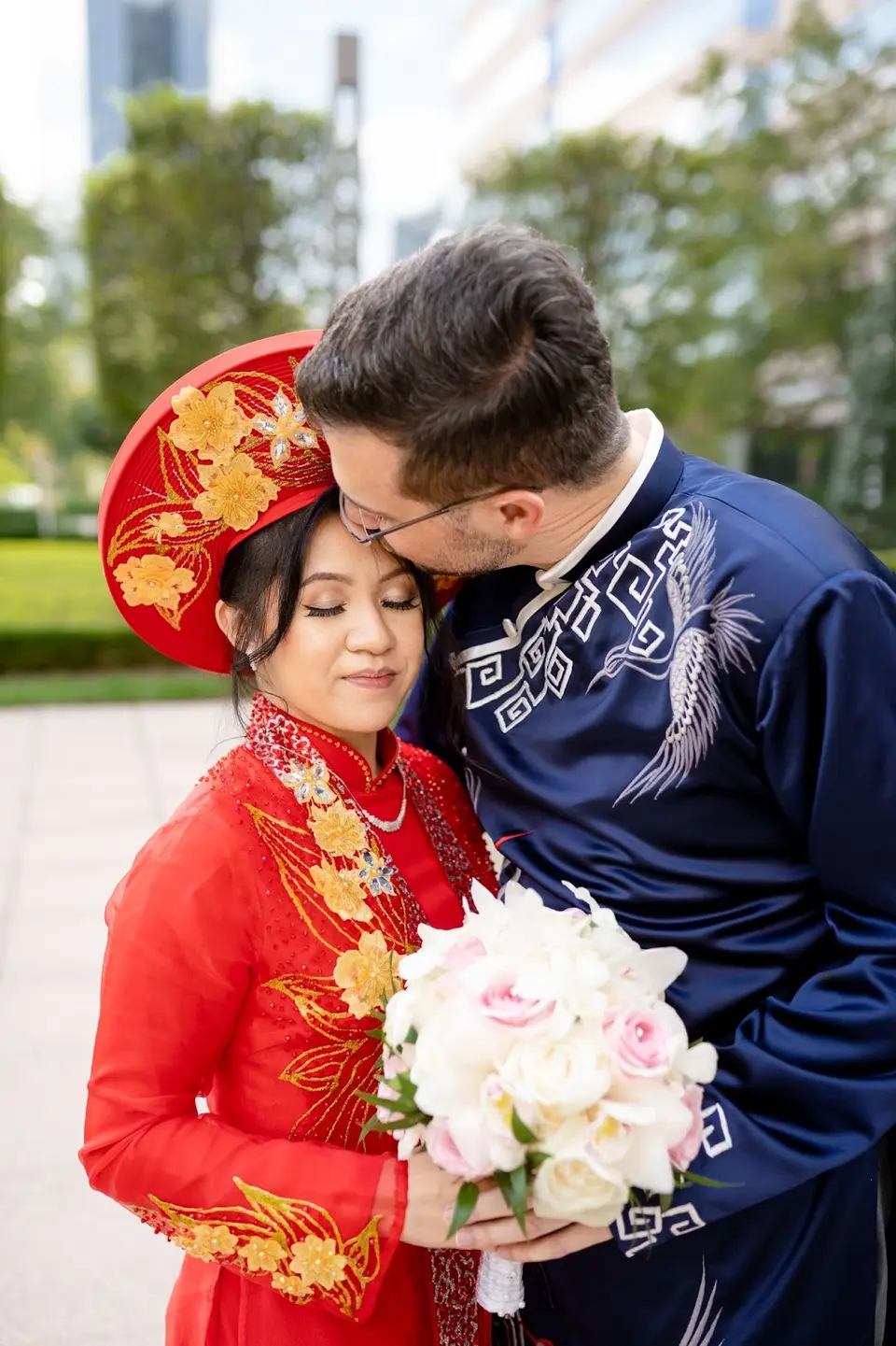 Bride in red ao dai with gold headpiece and groom in navy embroidered ao dai holding a bouquet