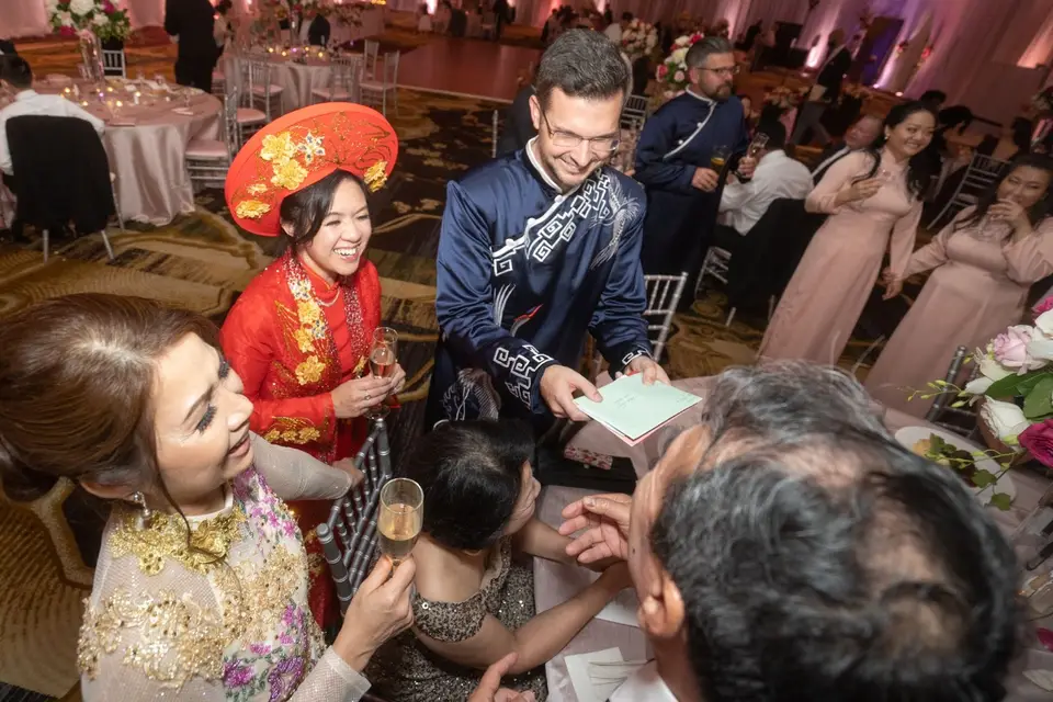 Groom in a navy embroidered ao dai handing out papers to seated guests beside the bride in red and gold