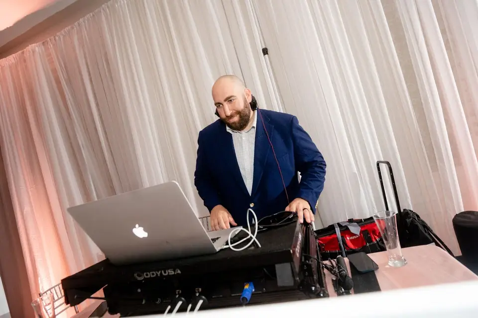 DJ in a navy blazer and headphones working at a laptop and controller behind draped curtains