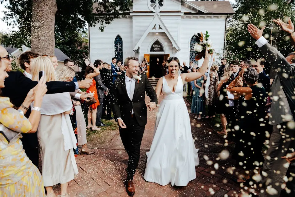 Bride and groom walking hand in hand out of a white chapel as guests toss confetti on a brick path