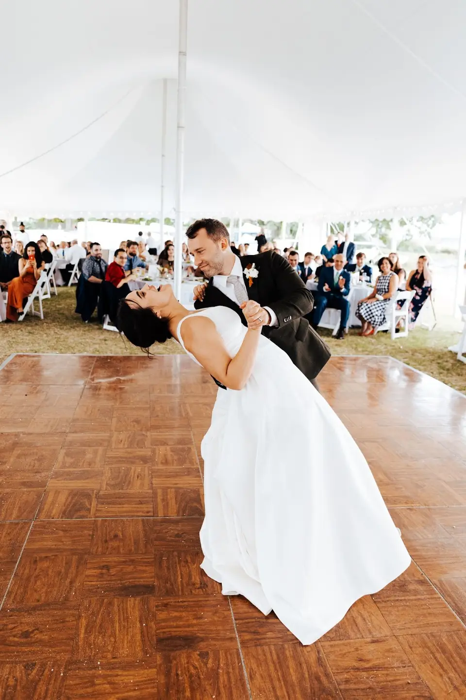 Groom dipping the bride during a first dance on a wood dance floor under a white tent with seated guests