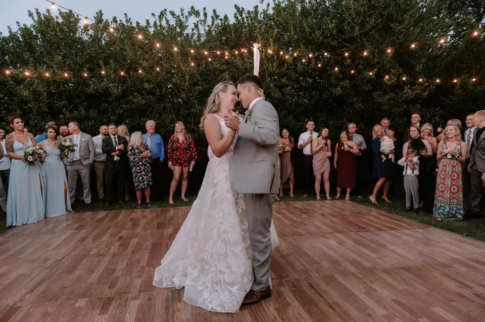 Bride and groom sharing a first dance on a wood floor under string lights as guests watch
