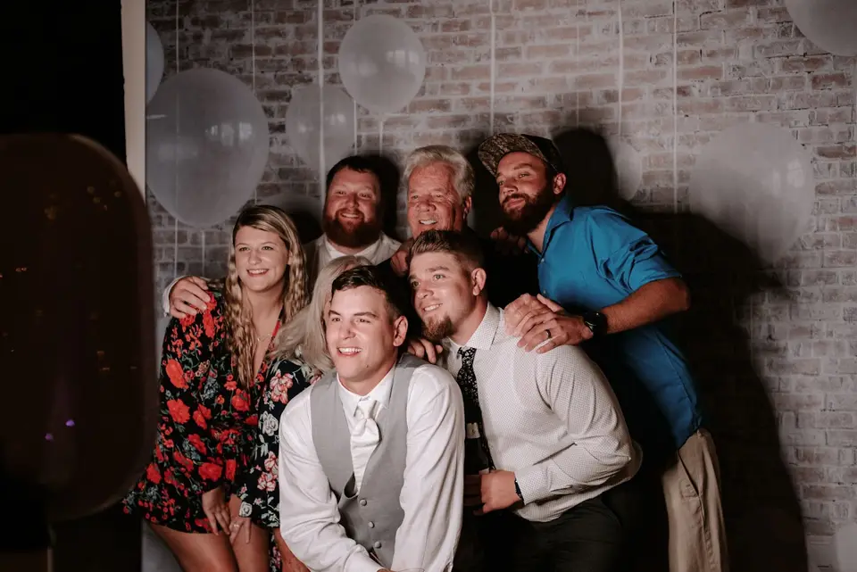 Group of friends posing close together in front of a white brick wall with balloons at a wedding photo booth