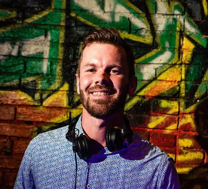 Smiling bearded man in a blue patterned shirt with headphones around his neck in front of graffiti