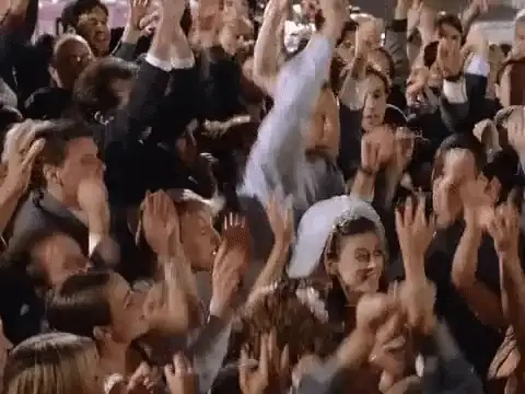 Movie still of a bride in a veil clapping among a cheering crowd of dancing guests