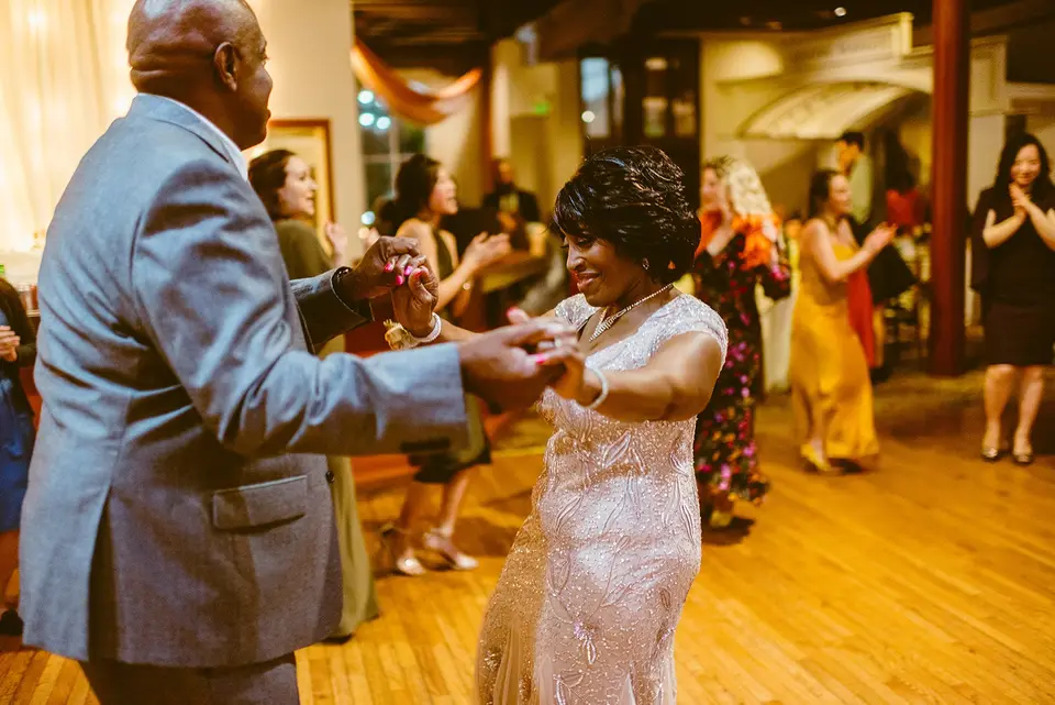 Older man in a gray suit dancing with a woman in a sparkling dress while guests dance behind them