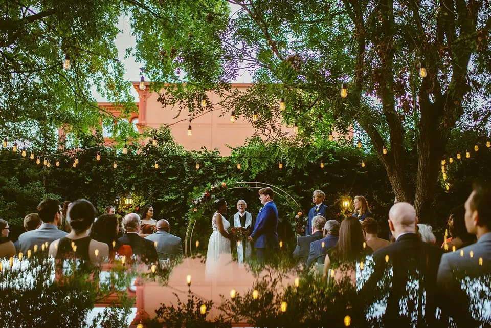 Outdoor wedding ceremony under an arch with guests seated beneath trees strung with lights