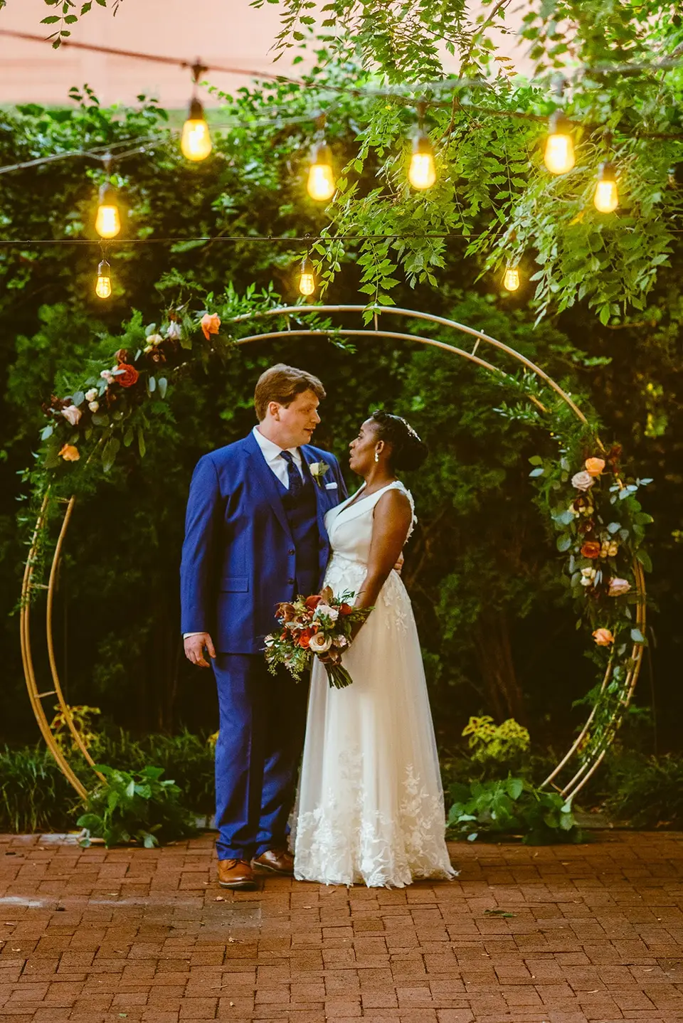 Bride and groom in a blue suit facing each other in front of a gold circular arch with flowers and string lights