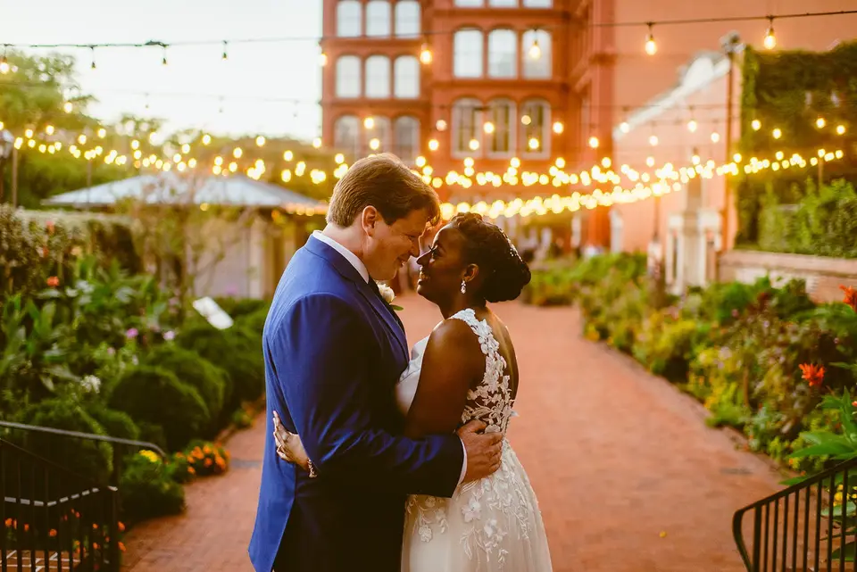 Bride and groom in a blue suit embracing on a brick path with string lights overhead