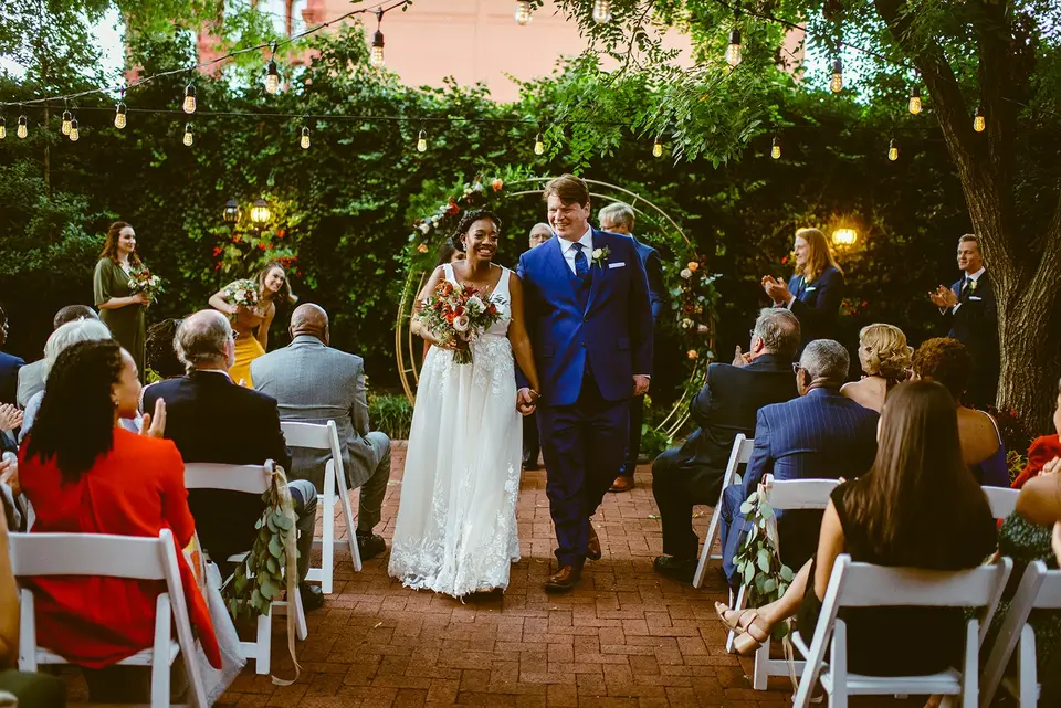 Bride and groom in a blue suit walking up the aisle between seated guests in an outdoor courtyard