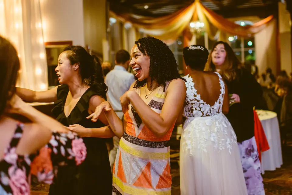 Smiling woman in an orange patterned dress dancing beside the bride and other guests at a reception