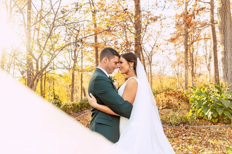 Bride and groom in a green suit embracing among autumn trees with her veil in the foreground