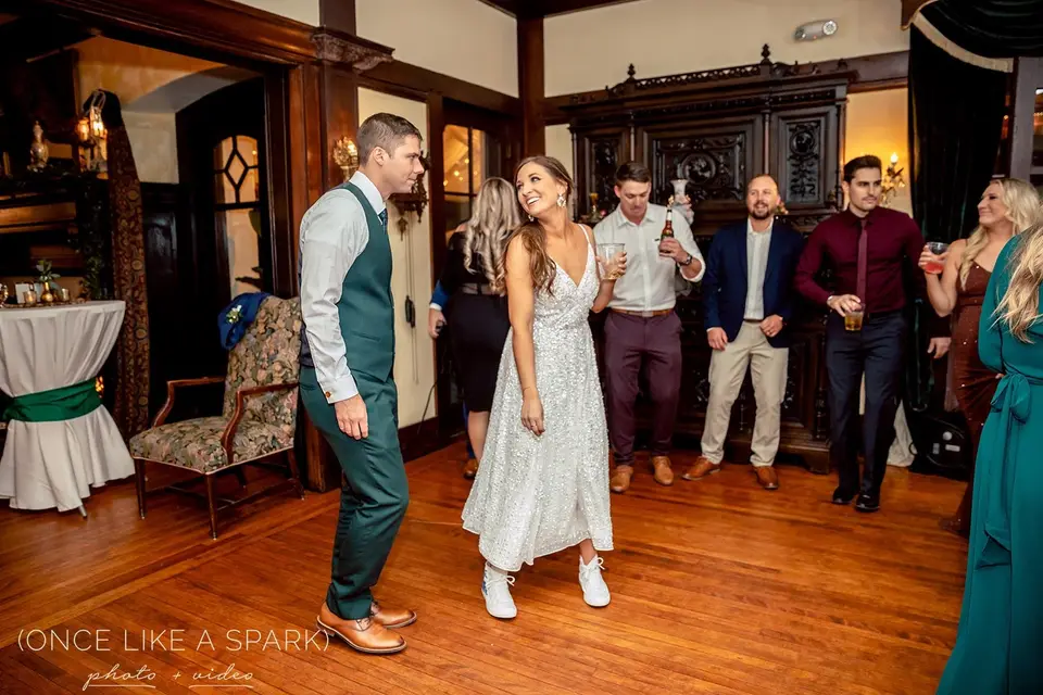 Groom in a green vest and bride in a sparkly dress stand among guests in a wood-paneled mansion room