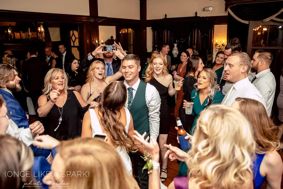 Crowded dance floor of guests dancing and filming on phones inside a wood-paneled room