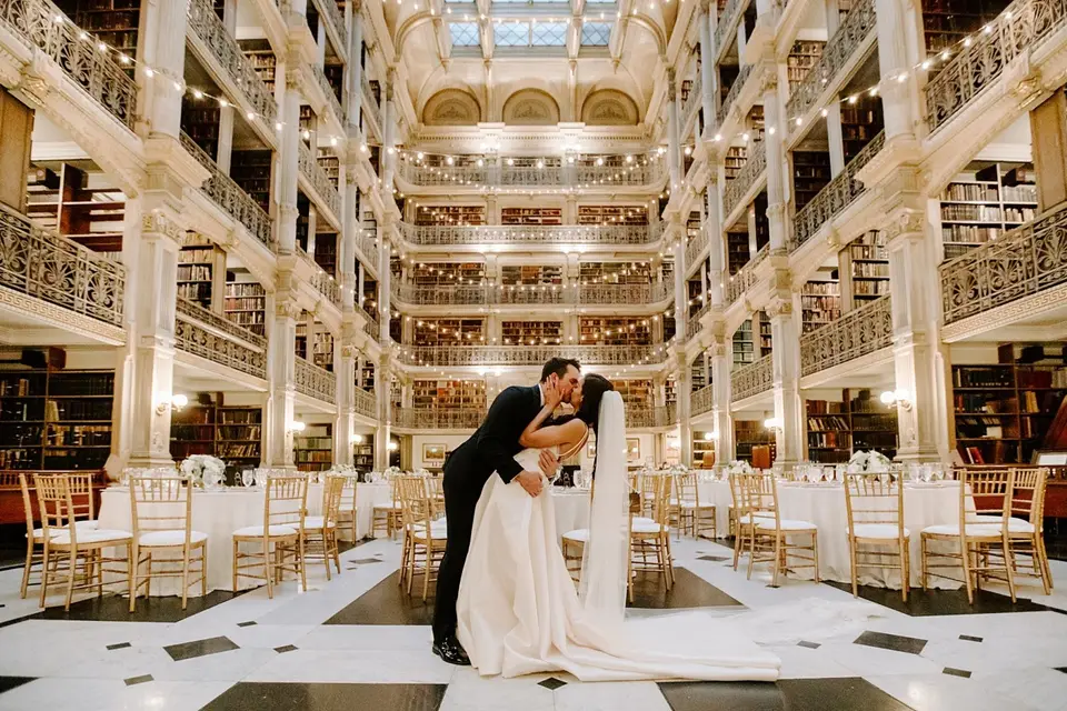 Bride and groom kissing in the middle of a grand multi-level library with gilded balconies and set tables